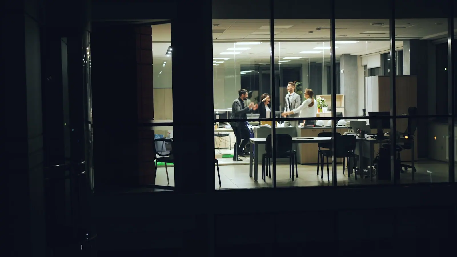 A team talking in a glass-walled office late in the evening, seen from outside